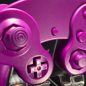 May include: Close-up of a purple games console controller. The controller has a directional pad, analogue stick, and buttons labelled A, B, and C. The design is a solid, vibrant purple, with a smooth, matte finish. The controller is designed for gaming.