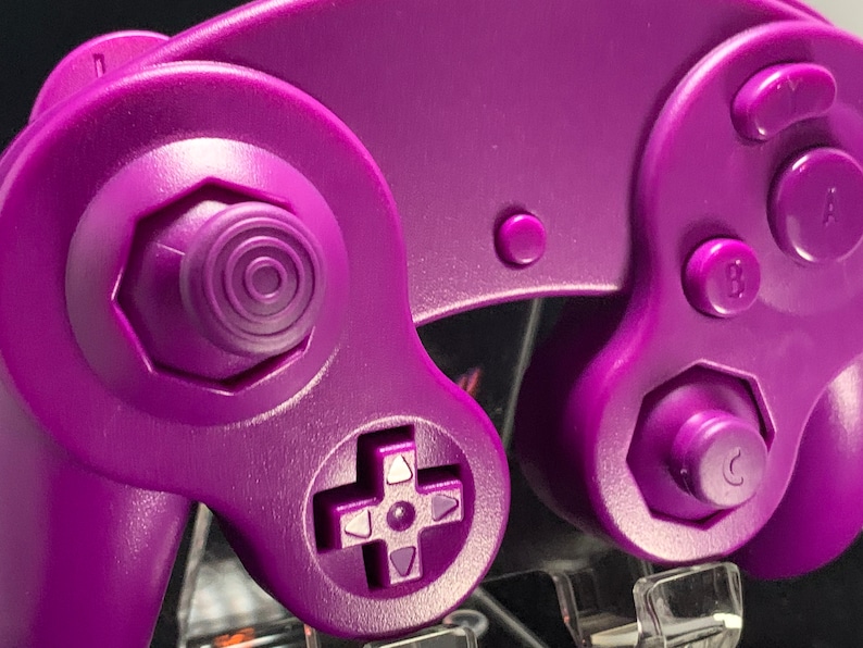 May include: Close-up of a purple games console controller. The controller has a directional pad, analogue stick, and buttons labelled A, B, and C. The design is a solid, vibrant purple, with a smooth, matte finish. The controller is designed for gaming.