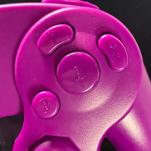May include: Close-up of a purple games console controller with the letters A, B, X, and Y on the buttons. The controller has a smooth, matte finish against a dark background.