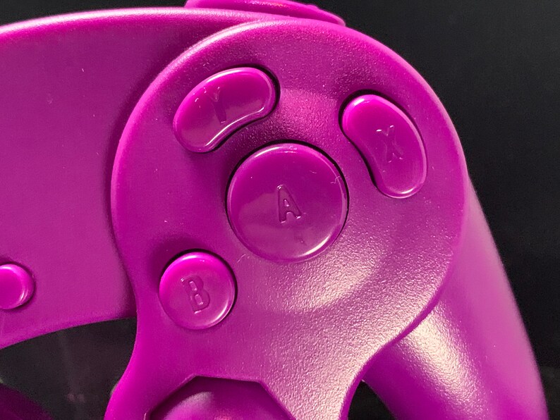 May include: Close-up of a purple games console controller with the letters A, B, X, and Y on the buttons. The controller has a smooth, matte finish against a dark background.