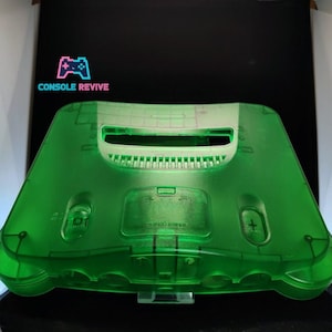May include: A transparent green Nintendo 64 console shell. The shell has a clear, see-through design, with visible internal components. The Console Revive logo is in the upper left corner. The console is displayed on a black surface.