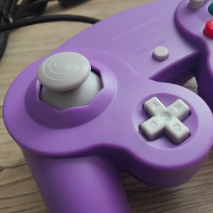 May include: Close-up of a purple video game controller with a gray joystick and white directional pad. The controller has red, yellow, and gray buttons. A black cord extends from the controller.