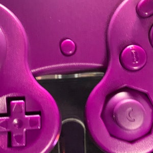 May include: Close-up of a purple video game controller. The controller features a directional pad, analog stick, and various buttons labelled A, B, and C. The design is a solid, vibrant purple, with a glossy finish. The image highlights the controller's ergonomic design.