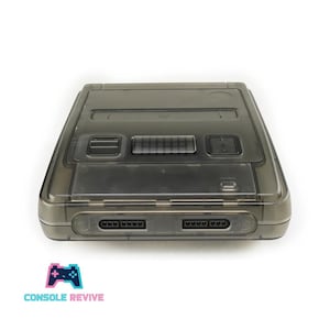 May include: A transparent gray Super Nintendo Entertainment System (SNES) console. The console has two controller ports, a power button, and a reset button. The text "CONSOLE REVIVE" is below the console.