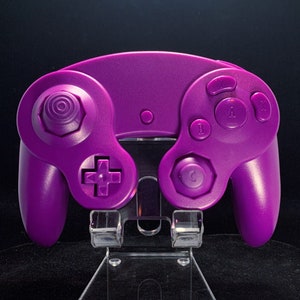 May include: A vivid purple games console controller. It has a distinctive shape with a directional pad, analogue stick, and buttons marked A, B, and C. The controller is displayed on a clear stand against a black backdrop.