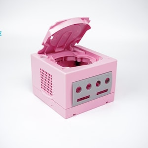 May include: A pink video game console with the top open, revealing the interior. The console has a gray faceplate with circular openings. The logo "CONSOLE REVIVE" is in the upper left corner.