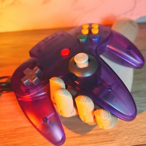 May include: A translucent purple video game controller with various coloured buttons, including yellow, green, blue, and red. The controller features a directional pad and an analogue stick. The controller rests on a wooden hand.
