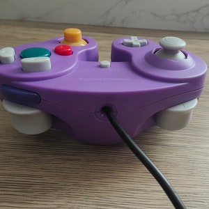 May include: A purple video game controller with a variety of colored buttons, including yellow, red, green, and gray. The controller has a gray joystick and a black cord.