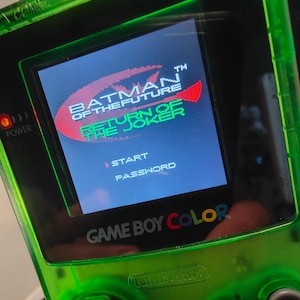 May include: A transparent green Game Boy Color handheld console showing the title screen for "Batman of the Future: Return of the Joker." The screen displays the game's logo in red, white, and green, with start and password options. The console's buttons are visible.