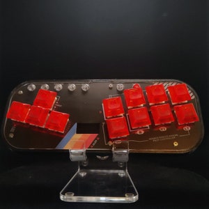 May include: A black and clear acrylic pilot controller with red buttons. The controller has a clear acrylic stand. The text "NOVACS REMOTE MANUAL PILOT CONTROLLER" is printed on the controller. The controller has a retro rainbow design.