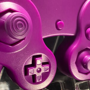 May include: Close-up of a purple video game controller. The controller features a directional pad, buttons labelled 'B' and 'C', and a joystick with concentric circles. The design is a solid, vibrant purple.