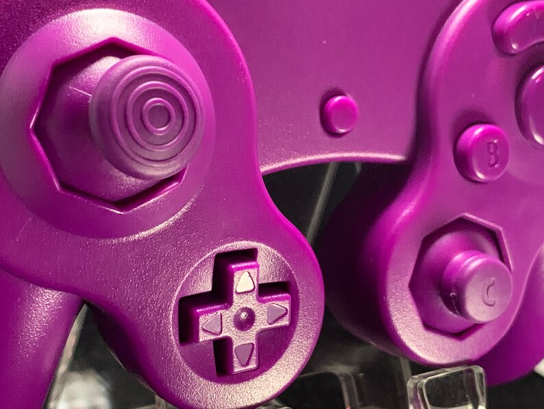 May include: Close-up of a purple video game controller. The controller features a directional pad, buttons labelled 'B' and 'C', and a joystick with concentric circles. The design is a solid, vibrant purple.