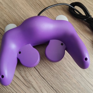 May include: A purple video game controller with a black cable and connector. The controller has two gray analog sticks and two purple buttons. The controller is designed for gaming.