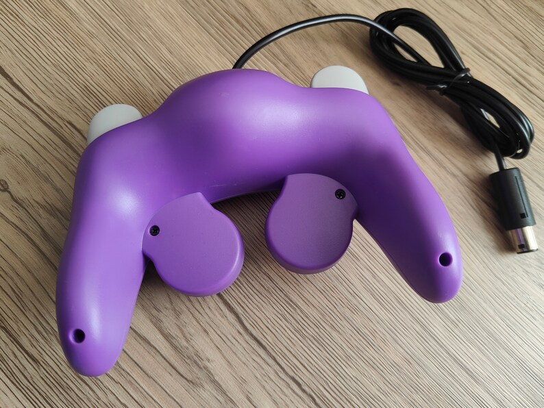 May include: A purple video game controller with a black cable and connector. The controller has two gray analog sticks and two purple buttons. The controller is designed for gaming.