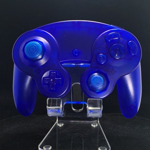 May include: A transparent blue video game controller on a clear plastic stand. The controller features a circular directional pad, a joystick, and various buttons. The stand has a rectangular base.