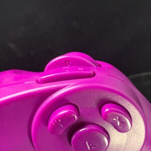 May include: Close-up of a purple video game controller. The controller features several buttons, including a directional pad, and buttons labeled with letters. The controller is made of smooth plastic.