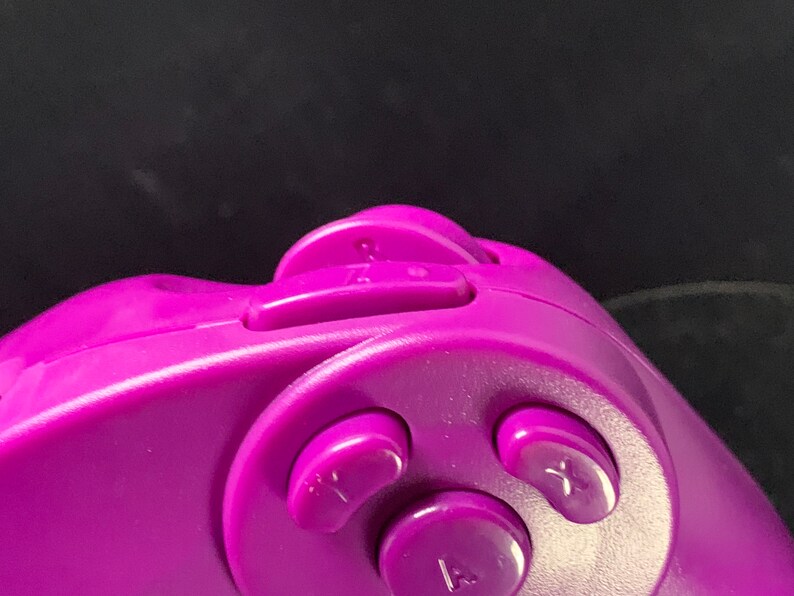 May include: Close-up of a purple video game controller. The controller features several buttons, including a directional pad, and buttons labeled with letters. The controller is made of smooth plastic.