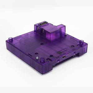 May include: A transparent purple electronic device with a rectangular base and a raised section on top. It has several ports and connectors visible on the sides and top. The device is made of a clear, glossy plastic.