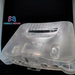 May include: A transparent Nintendo 64 console shell. The shell is rectangular with curved edges, and has openings for buttons and ports. The word "RESET" is visible on the front. The logo "CONSOLE REVIVE" is in the upper left corner.