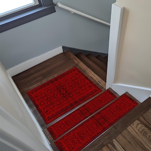 Może przedstawiać: Czerwone stopnie schodów z czarnym, ozdobnym wzorem umieszczone na drewnianych schodach. Stopnie są prostokątne i zakrywają szerokość stopni. Schody znajdują się w domu z szarymi ścianami i białą poręczą.