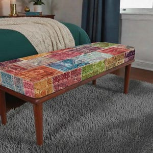 May include: A rectangular bench with a patchwork design seat in shades of red, orange, blue, and green. The bench has a dark brown wooden frame and four tapered legs. It is placed on a gray, shaggy rug, with a bed and window in the background.