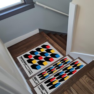 Può includere: Tappeti per gradini con un design geometrico in rosso, giallo, blu e nero. I tappeti sono rettangolari e presentano il testo "Bauhaus Ausstellung Jul. August. September 1923". I tappeti sono posizionati su scale di legno.