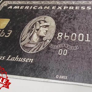 May include: A black American Express credit card with a gold embossed logo and the text "AMERICAN EXPRESS" in white. The card number is 3763 86001 and the name "Dr. Nikolaus Lahusen" is printed below the logo. The card has a silver border and the text "MEMBER SINCE 00" is printed below the card number.