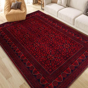 Puede incluir: Una alfombra rectangular roja con un patrón geométrico detallado y un borde azul marino oscuro. La alfombra presenta diseños intrincados. Se encuentra sobre un suelo de madera clara, con un sofá blanco y un puf beige en el fondo.