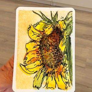 Könnte beinhalten: Ein rechteckiges Aquarellbild auf Papier, das eine leuchtend gelbe Sonnenblume mit einer dunkelbraunen, strukturierten Mitte, grünen Blättern und schwarzen Tintenumrissen zeigt. Das Kunstwerk ist auf hellgelbem Grund und trägt die Initialen mj in schwarzer Tinte.