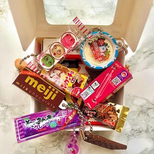 May include: A selection of Japanese sweets and snacks in an open white box. Items include Meiji milk chocolate, colourful candy packages, and a keychain with a cartoon character. The box is on a marble-patterned surface.