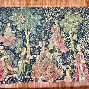 Puede incluir: Un tapiz que representa una escena de un jardín medieval con figuras con ropa colorida. El tapiz presenta un gran árbol con pájaros, flores y una variedad de follaje. Las figuras están dispuestas en grupo, algunas de pie y otras sentadas. El tapiz está tejido con intrincados detalles y colores vibrantes.