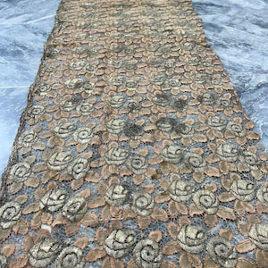 May include: A close-up of a piece of lace fabric with a delicate floral pattern. The lace is a combination of gold and brown colours, creating a vintage and elegant look.