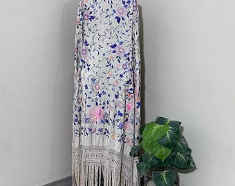 May include: A long, cream-coloured shawl with a floral pattern in pink, purple, and blue. The shawl has a fringed bottom and is draped over a form. A potted plant with large green leaves is in the corner.