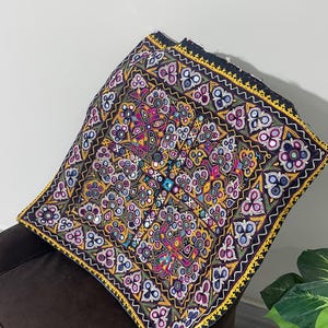Op de afbeelding: Vierkante, geborduurde stof met een zwarte achtergrond en een levendig bloemenpatroon. Het ontwerp bevat meerdere kleuren, waaronder roze, geel, blauw en groen, met spiegelaccenten. De randen zijn afgewerkt met gele en zwarte details.