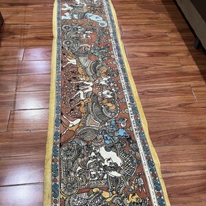 Puede incluir: Un tapiz de pared de tela largo y estrecho pintado a mano con una representación detallada de una procesión de figuras y animales. Las figuras visten trajes elaborados y montan a caballo y elefante. El fondo es de un color marrón intenso con acentos amarillos y azules.