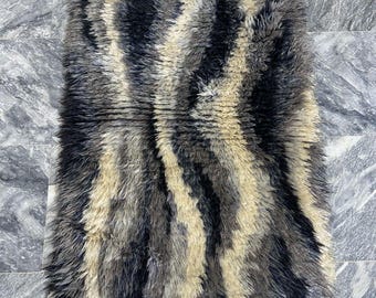 May include: Rectangular rug with a textured, shaggy pile. The rug features vertical stripes in shades of black, gray, and beige. The edges are finished with a fringe. This rug is suitable for a living room or bedroom.