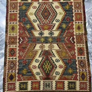 Può includere: Un tappeto tradizionale con un motivo geometrico in tonalità di rosso, marrone, beige e blu. Il tappeto ha un bordo con frange.