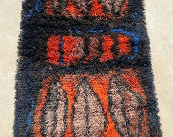 May include: A vintage rug with a geometric pattern in shades of orange, brown, blue, and black. The rug has a shaggy texture and a fringe border.
