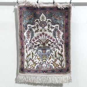 May include: A rectangular, ornate rug with a cream-colored background and a detailed floral and bird design. The rug features a central vase with flowers, surrounded by birds and other decorative elements. The border is a deep burgundy color, and the rug has a fringe at the bottom.