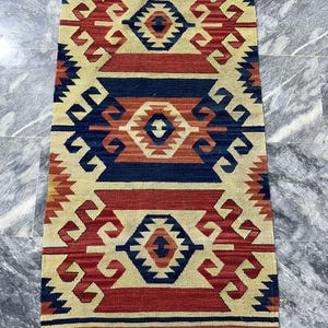 May include: A rectangular rug with a geometric pattern in red, navy blue, and beige. The rug features repeating diamond and angular shapes. The edges have a fringe. This rug is suitable for a living room or bedroom.