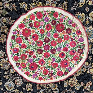 May include: A white circular tablecloth with a red, pink, green, and blue floral embroidery pattern. The tablecloth is on a black background with a floral pattern.
