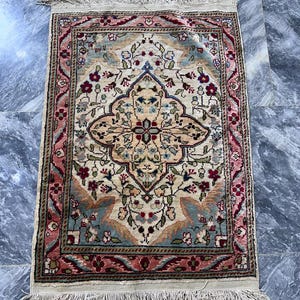 May include: A rectangular rug with a cream-colored central design featuring floral and geometric patterns in shades of red, blue, and green. The rug has a red border and cream fringe along the edges. The rug measures approximately 24 inches long and 16 inches wide.