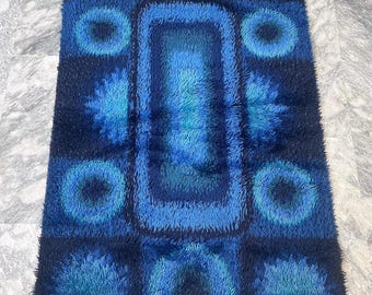 Puede incluir: Una alfombra de pelo azul y negro con un patrón geométrico de círculos y cuadrados. La alfombra tiene una pila alta y una textura suave.