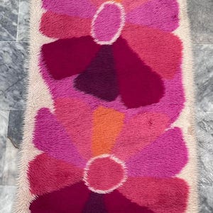 May include: A rectangular rug with a floral design. The rug features two large flowers with petals in shades of pink, fuchsia, and orange. The flowers have a white center and are set against a beige background.