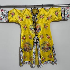 May include: A yellow silk robe with intricate floral and landscape embroidery. The robe has a mandarin collar and long sleeves with a decorative trim.