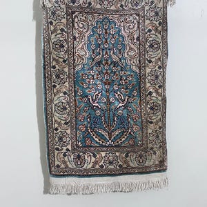 May include: A small, rectangular rug with a central teal design featuring a tree and birds. The rug has a cream-colored border with floral patterns and fringe along the bottom edge. The rug is hanging from a metal rod.