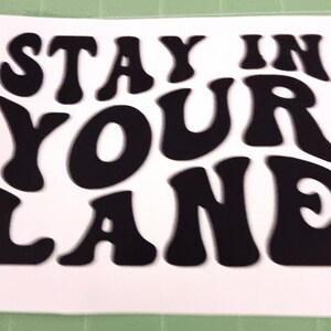 Könnte beinhalten: Ein weißer Aufkleber mit dem Text "STAY IN YOUR LANE" in fetter, schwarzer Retro-Schrift. Die Schriftart ist abgerundet, was eine Vintage-Ästhetik suggeriert. Geeignet für verschiedene Oberflächen.