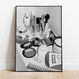 Op de afbeelding: Zwart-witfoto van een verzameling schoonheidsproducten en accessoires. De afbeelding bevat make-up, parfumflessen, een kam, oorbellen en een kaars. Een CHANEL-compact is zichtbaar.