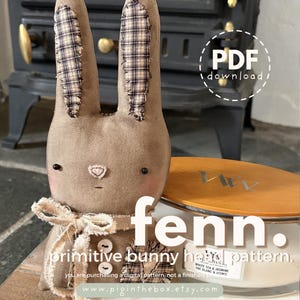 May include: A handmade brown fabric bunny with plaid ears and a bow tie. The bunny has button accents and a stitched face. Text on the image reads "fenn. primitive bunny head pattern." A PDF download icon is visible.