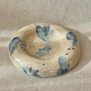 Könnte beinhalten: Handgefertigte Keramikschale mit kreisförmigem Design. Die Schale ist cremefarben mit abstrakten blauen Mustern und Sprenkeln. Der Rand ist leicht erhöht und die Schale hat eine glänzende Oberfläche. Geeignet zum Servieren oder Dekorieren.
